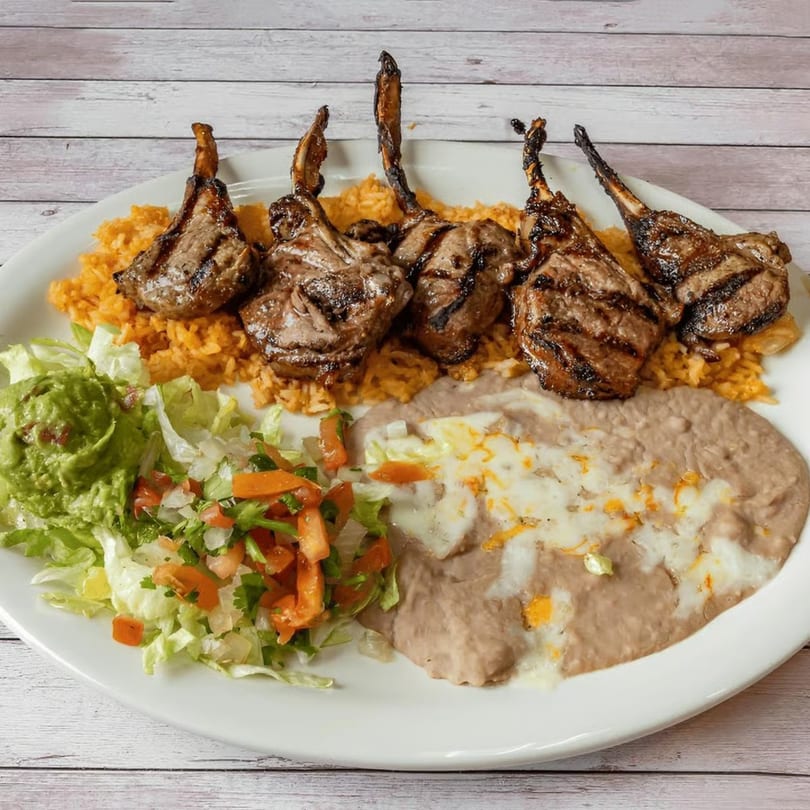 Grilled lamb chops with rice, refried beans, guacamole, and a side salad on a white plate.