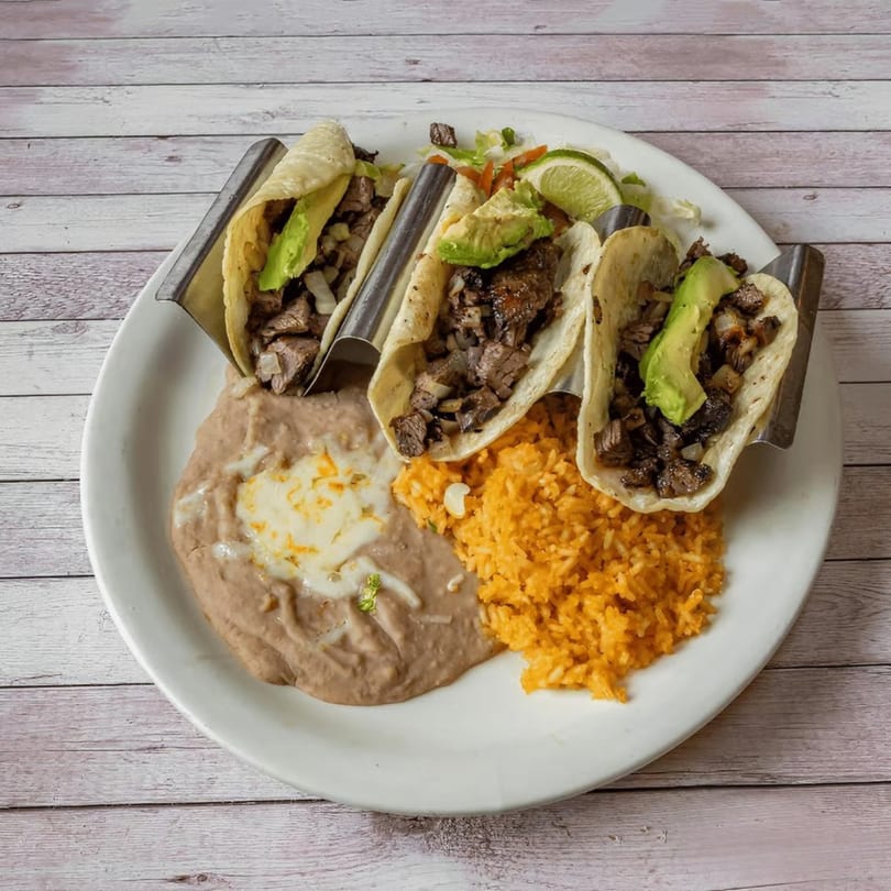 Three steak tacos with avocado, refried beans with cheese, and orange rice on a white plate.