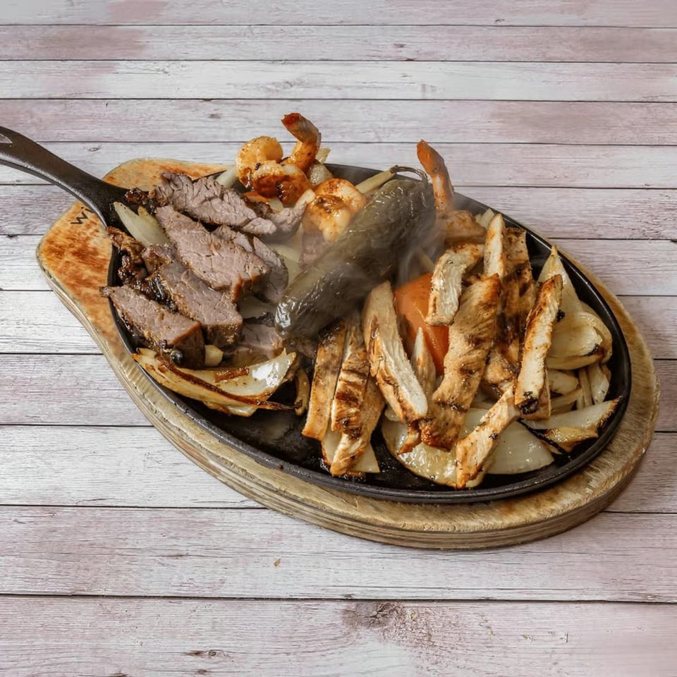 Sizzling fajitas with steak, chicken, shrimp, onions, and peppers in a cast iron skillet.