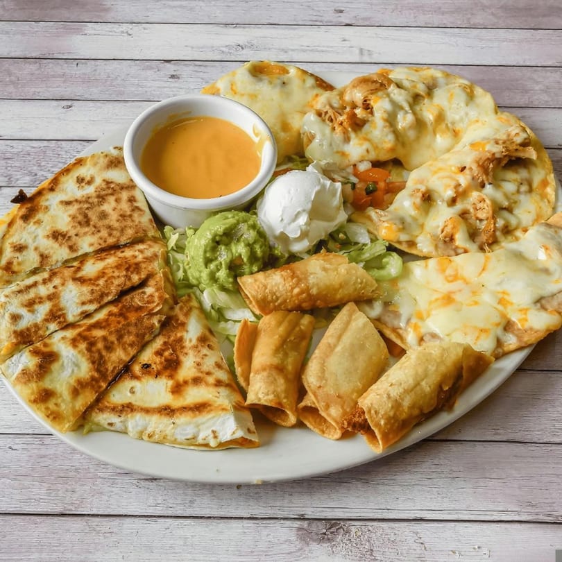 A large platter of Mexican appetizers including quesadillas, taquitos, nachos, guacamole, sour cream, and cheese dip.